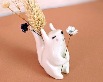 May include: White ceramic squirrel figurine with a closed eye, holding a single white flower. The squirrel is decorated with black painted eyes and a small mouth. The figurine is holding a single white flower with a green stem.
