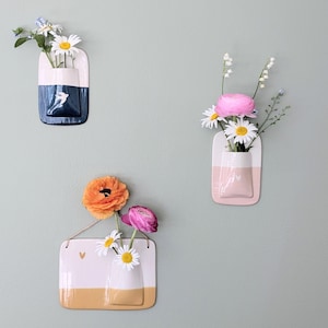 Peut inclure: Trois vases muraux en céramique, chacun contenant des fleurs fraîches. Un vase est bleu et blanc, un autre rose et blanc, et le troisième blanc et jaune. Les vases sont rectangulaires et ont une forme de poche pour contenir les fleurs.