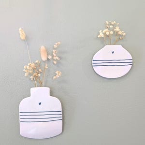 Peut inclure: Deux vases muraux en céramique blanche avec des fleurs séchées. Chaque vase a un cœur bleu et des rayures bleues horizontales. Un vase est rectangulaire, l'autre est rond. Les vases sont montés sur un mur vert clair.