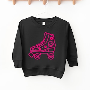 以下が含まれることがあります： 花と笑顔のついたローラースケートのピンク色のアウトラインが描かれた黒いスウェットシャツ