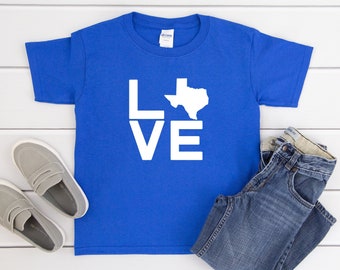 May include: A royal blue short-sleeved t-shirt featuring the white text LOVE, where the letter O is replaced by a white silhouette of the state of Texas. The shirt is displayed alongside a pair of gray slip-on shoes and blue denim jeans.