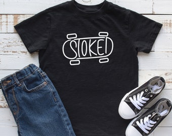 Może przedstawiać: Czarny t-shirt z białym napisem "STOKED" w kształcie deskorolki. W zestawie z niebieskimi dżinsowymi szortami i czarno-białymi trampkami. Przedmioty ułożone są na białej drewnianej powierzchni.
