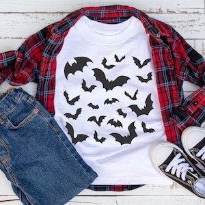 May include: A white t-shirt with a black bat pattern. The shirt is laid out on a white wooden surface with a red and black plaid shirt, blue jeans, and black sneakers.