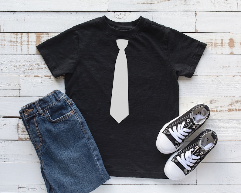 Op de afbeelding: Een zwart T-shirt met een witte stropdas-afbeelding. Het shirt ligt op een wit houten oppervlak met een paar zwart-witte sneakers en een blauwe jeans.