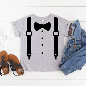 May include: A gray t-shirt with a black bow tie and suspenders design. The shirt is laid flat on a white wooden surface with a denim jacket, a white headband, and two pairs of sandals.