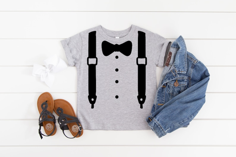 May include: A gray t-shirt with a black bow tie and suspenders design. The shirt is laid flat on a white wooden surface with a denim jacket, a white headband, and two pairs of sandals.