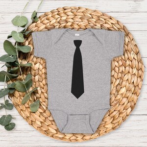Op de afbeelding: Een grijze baby romper met een zwarte stropdas afbeelding op de voorkant.