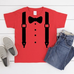 May include: Red t-shirt with a black bow tie and suspenders graphic. The shirt is on a white wood background with a pair of gray loafers and blue jeans.