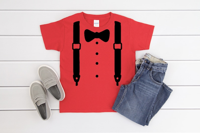 May include: Red t-shirt with a black bow tie and suspenders graphic. The shirt is on a white wood background with a pair of gray loafers and blue jeans.