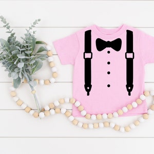 May include: A pink baby onesie with a black bow tie and suspenders graphic.