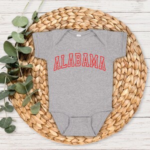 May include: A gray baby onesie with red text that reads "ALABAMA". The onesie is on a woven straw mat.