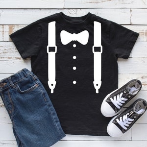 May include: Black t-shirt with a white bow tie and suspenders design. The shirt is laid flat on a white wooden surface with a pair of black canvas shoes and blue jeans.