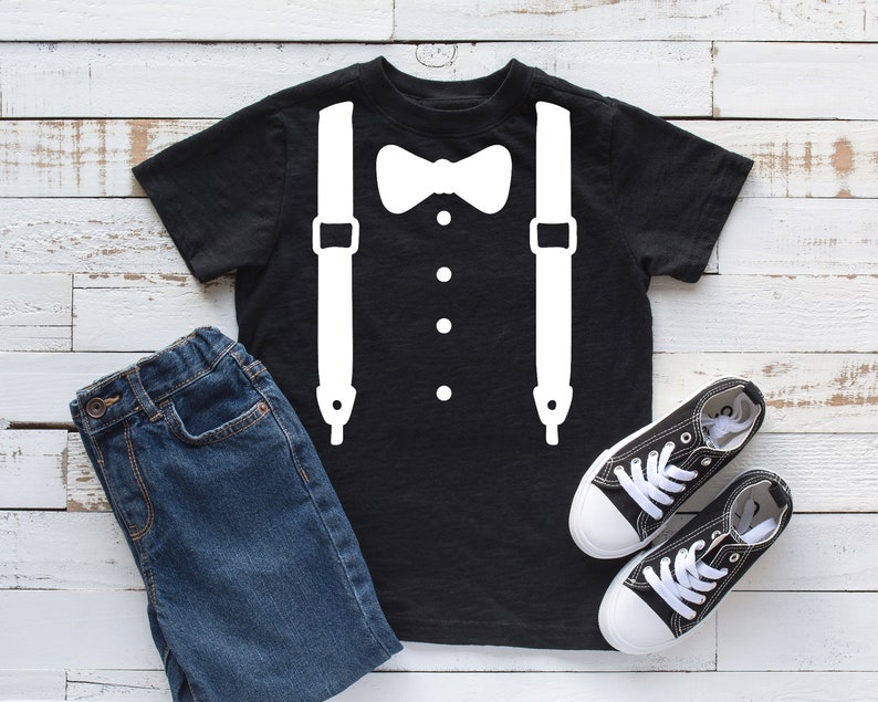 May include: Black t-shirt with a white bow tie and suspenders design. The shirt is laid flat on a white wooden surface with a pair of black canvas shoes and blue jeans.