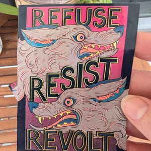 May include: A rectangular sticker with the words "REFUSE, RESIST, REVOLT" in bold, outlined letters. The sticker features two stylized wolf illustrations with snarling expressions, in shades of gray, pink, and blue.