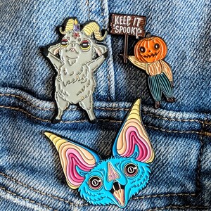 May include: Three enamel pins on denim. One is a gray goat with a pentagram, another is a pumpkin-headed scarecrow holding a sign that says "Keep It Spooky", and the last is a blue bat with pink and yellow accents.