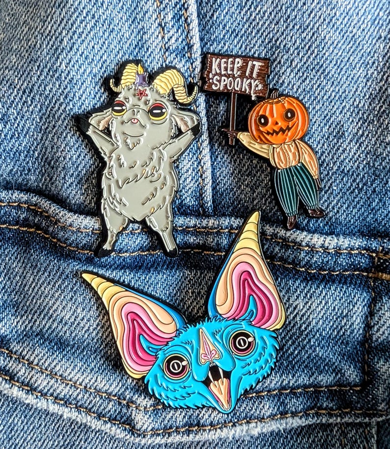 May include: Three enamel pins on denim. One is a gray goat with a pentagram, another is a pumpkin-headed scarecrow holding a sign that says "Keep It Spooky", and the last is a blue bat with pink and yellow accents.
