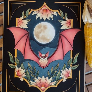 May include: A digital illustration of a bat with pink wings flying in front of a full moon. The bat is surrounded by white flowers and green leaves on a dark blue background. The image is framed with a gold and black border.