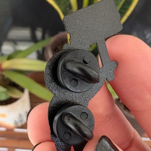 May include: Black enamel pin depicting a figure holding a sign. The pin has two black pin backs. The figure is holding a sign with a textured surface. The pin is a unique accessory.