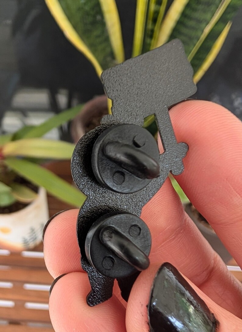 May include: Black enamel pin depicting a figure holding a sign. The pin has two black pin backs. The figure is holding a sign with a textured surface. The pin is a unique accessory.