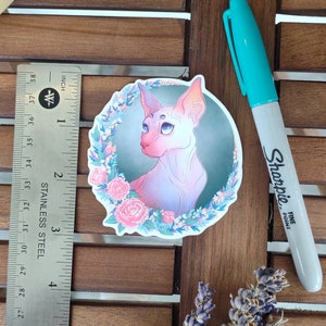 以下が含まれることがあります： 青い目をしたスフィンクス猫のカラフルなイラストが描かれたステッカー。ピンクと紫の花に囲まれています。猫は濃い緑色の背景に描かれています。