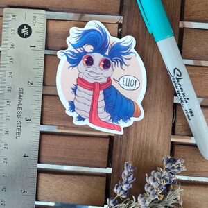 以下が含まれることがあります： 赤いスカーフをした青と白の漫画の虫で、吹き出しに「Ello！」と書かれています。虫は大きな目をしていて、友好的な表情をしています。ステッカーは木製の表面に貼られています。