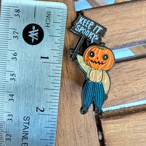 May include: A Halloween-themed enamel pin featuring a smiling jack-o'-lantern holding a sign that reads "Keep It Spooky." The pin is approximately 1 inch (2.5 cm) tall and has a light brown shirt, blue pants, and black shoes.