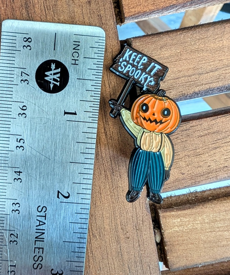 May include: A Halloween-themed enamel pin featuring a smiling jack-o'-lantern holding a sign that reads "Keep It Spooky." The pin is approximately 1 inch (2.5 cm) tall and has a light brown shirt, blue pants, and black shoes.