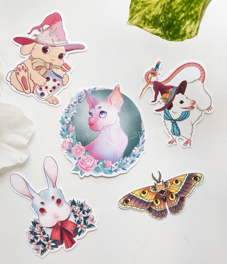 以下が含まれることがあります： 幻想的な動物のイラストが描かれた、カラフルなステッカー5枚。赤い蝶ネクタイをした白いウサギ、ピンクの魔女の帽子をかぶった白いウサギ、青いスカーフと魔女の帽子をかぶった白いネズミ、ピンクの目をした白い毛のない猫、そして大きな目をした茶色と黄色の蛾。