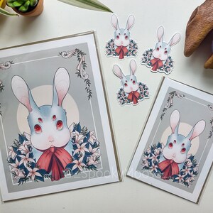 May include: A set of four prints featuring a white rabbit with red eyes and a red bow. The rabbit is surrounded by white flowers and green leaves. The prints are on a white background with a gray border.