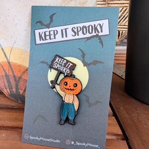 May include: A Halloween-themed enamel pin featuring a pumpkin-headed character holding a sign that reads "KEEP IT SPOOKY." The pin is set against a blue background with bats and a full moon. The pin is attached to a card with the text "KEEP IT SPOOKY."