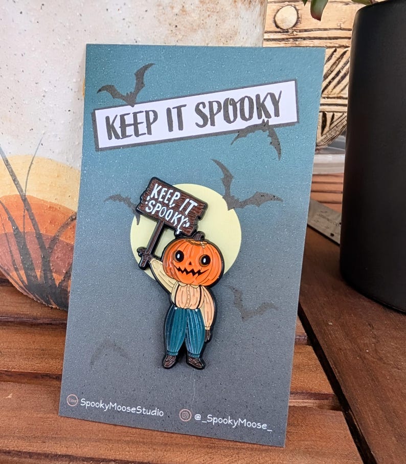 May include: A Halloween-themed enamel pin featuring a pumpkin-headed character holding a sign that reads "KEEP IT SPOOKY." The pin is set against a blue background with bats and a full moon. The pin is attached to a card with the text "KEEP IT SPOOKY."