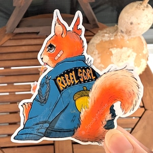 May include: A sticker of a cartoon squirrel wearing a blue denim jacket with the words "Rebel Squirrel" on the back. The squirrel has a bandage on its eye and a brown acorn on its jacket.