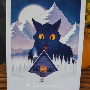 May include: A digital illustration of a large black cat with glowing yellow eyes standing in front of a small wooden cabin in a snowy mountain landscape. The cat is wearing a red ribbon around its neck with a bell attached. The moon is visible in the night sky.