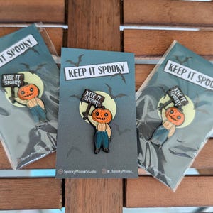 May include: Three Halloween-themed enamel pins featuring a pumpkin-headed character holding a sign that reads "KEEP IT SPOOKY." The pins are on cards with a dark blue background, bats, and a full moon. The cards are in clear plastic packaging.