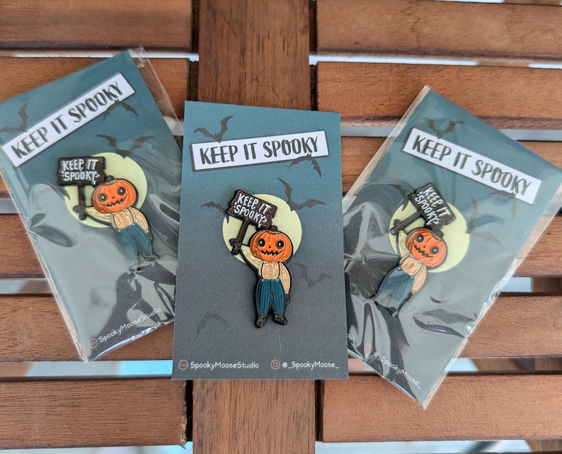 May include: Three Halloween-themed enamel pins featuring a pumpkin-headed character holding a sign that reads "KEEP IT SPOOKY." The pins are on cards with a dark blue background, bats, and a full moon. The cards are in clear plastic packaging.