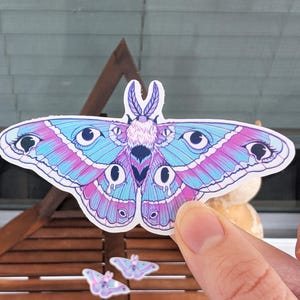 Puede incluir: Una pegatina de una polilla azul y rosa con ojos grandes. La polilla tiene un cuerpo peludo y patrones intrincados en las alas.
