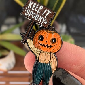 May include: A Halloween-themed enamel pin featuring a pumpkin-headed character holding a sign that reads "KEEP IT SPOOKY." The character wears a tan shirt, teal pants, and black shoes. The sign is brown with white lettering.