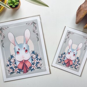 May include: A print of a white rabbit with four eyes and a red bow tie. The rabbit is surrounded by white flowers and green leaves. The background is a light gray with a white circle in the center.