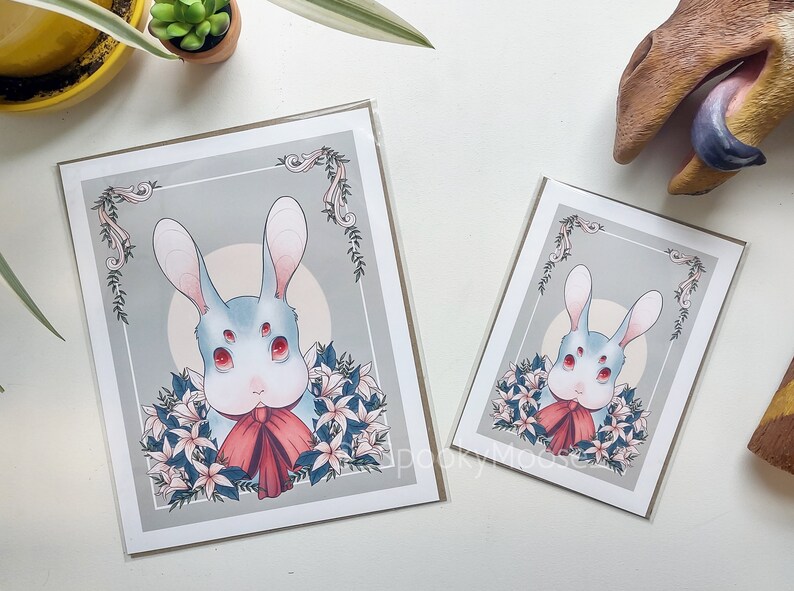 May include: A print of a white rabbit with four eyes and a red bow tie. The rabbit is surrounded by white flowers and green leaves. The background is a light gray with a white circle in the center.