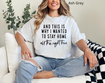 May include: A woman wearing a light gray t-shirt with the text "AND THIS IS WHY I WANTED TO STAY HOME all this, right here." The shirt is a relaxed fit and has a crew neck. The woman is sitting on a white couch with a black and white checkered throw blanket.