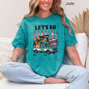 May include: Jade green t-shirt with the text "LET'S GO HOKING" above a graphic of a truck and a cartoon dog. The truck is white and green. The text "San Antonio" is also visible.