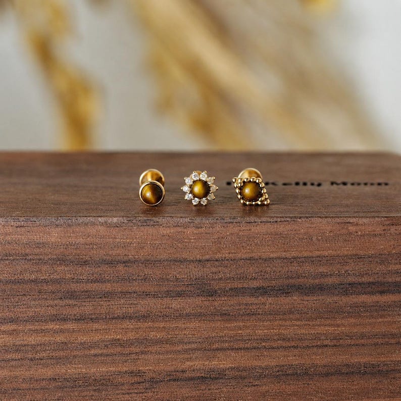 May include: Three gold stud earrings with tiger's eye stones. One earring is a simple round stud, another features a flower design with small clear stones, and the third has a beaded edge.