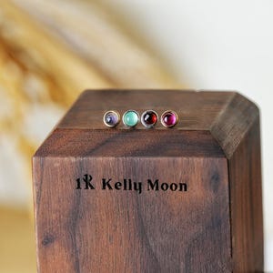 以下が含まれることがあります： 紫、ターコイズ、濃い赤、ピンクのカラフルな宝石が付いた4つのスタッドピアス。各宝石は金色のベゼルにセットされています。木製のブロックには「1R Kelly Moon」の文字が表示されています。