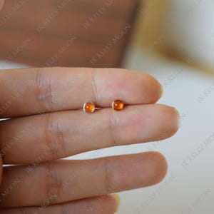 May include: A pair of small stud earrings with round, orange-colored gemstones set in silver-colored metal. The earrings are displayed on a light-colored background, showcasing their simple design and warm color.
