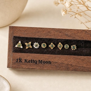 May include: A set of eight gold-toned stud earrings with green moss agate stones, displayed in a dark brown wooden box with the text Kelly Moon engraved on the front. The earrings feature various geometric shapes, including star, floral, and circular designs.