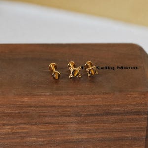 May include: Three gold-colored stud earrings with tiger's eye stones. One earring is a simple round stud, while the other two have decorative gold settings. The earrings are displayed on a wooden surface. The text "Kelly Moon" is visible.