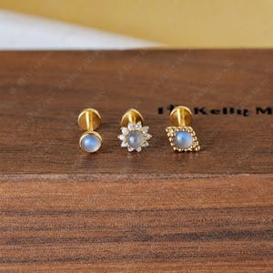 Puede incluir: Tres pendientes de botón dorados con cabujones de piedra lunar iridiscente. Uno es redondo, otro tiene forma de flor y otro tiene forma de diamante con un borde de cuentas. Los pendientes se muestran sobre una superficie de madera.
