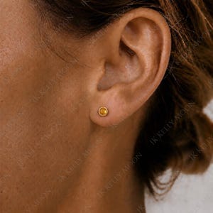 May include: A small, round stud earring featuring a single, faceted yellow gemstone set in a polished gold-toned metal bezel. The earring is positioned centrally on the earlobe, showcasing a minimalist design with a warm, golden hue.