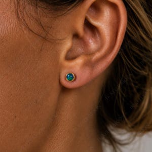 May include: A small, round stud earring featuring a bezel-set, iridescent blue and green opal stone encased in a polished gold-tone metal setting, securely positioned in the center of an earlobe, showcasing a vibrant play of color.