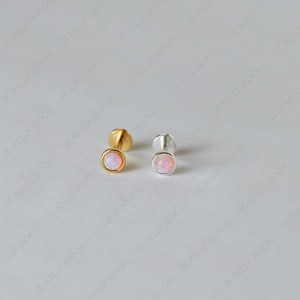 May include: Two stud earrings with round, light pink opal stones. One earring has a gold setting, and the other has a silver setting. The earrings are displayed on a light background.